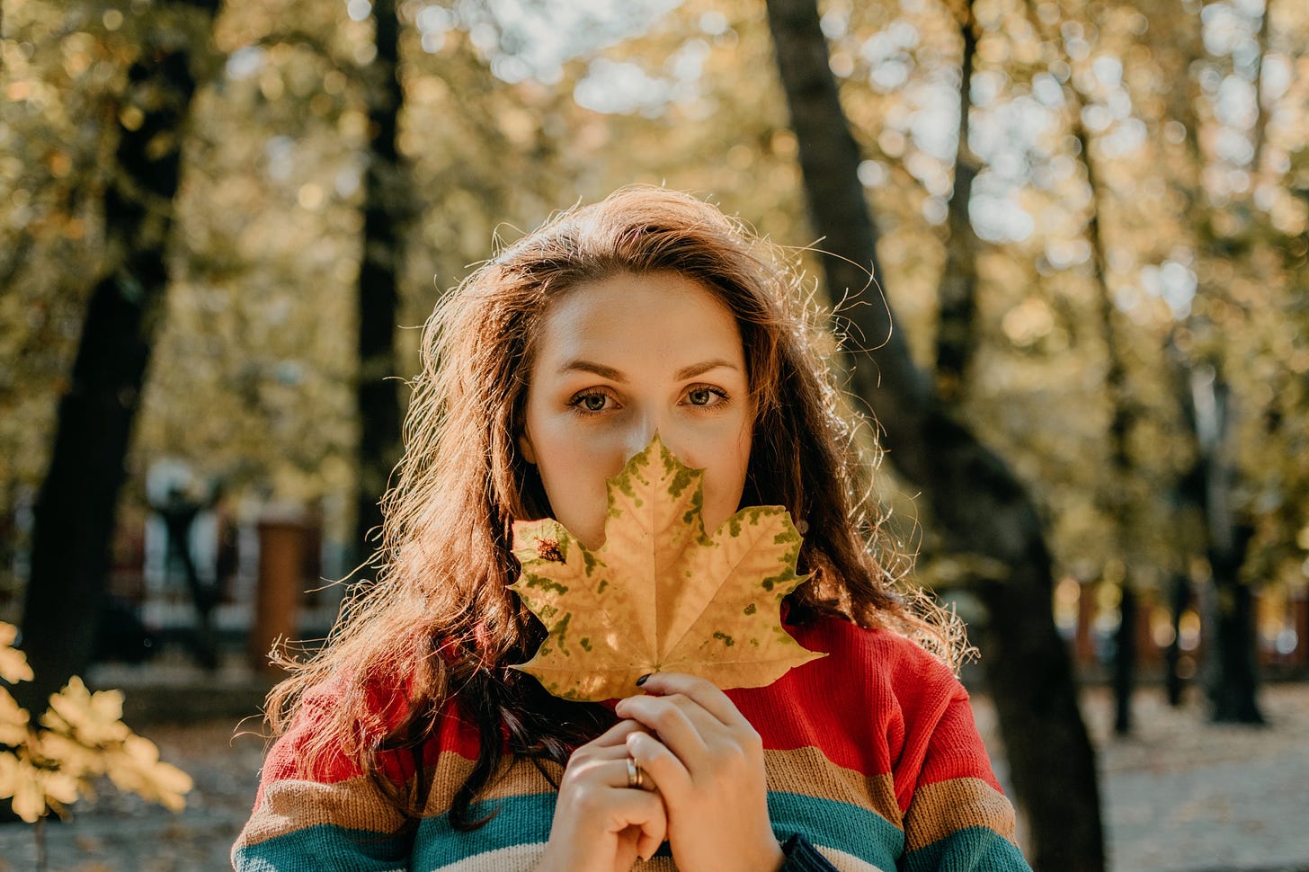 A woman conceals her face behind a large, yellowing leaf in an autumn park.