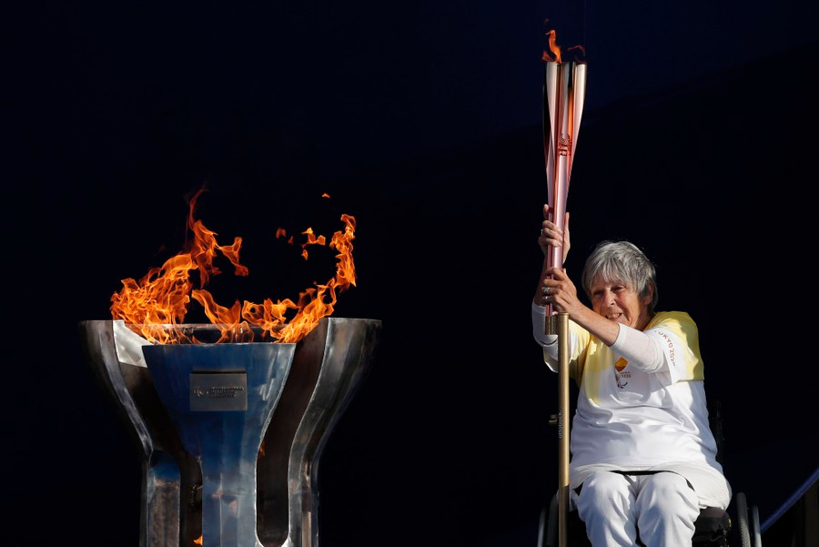 A woman in a wheelchair holds a lit torch beside a metal cauldron with flames rising above it.