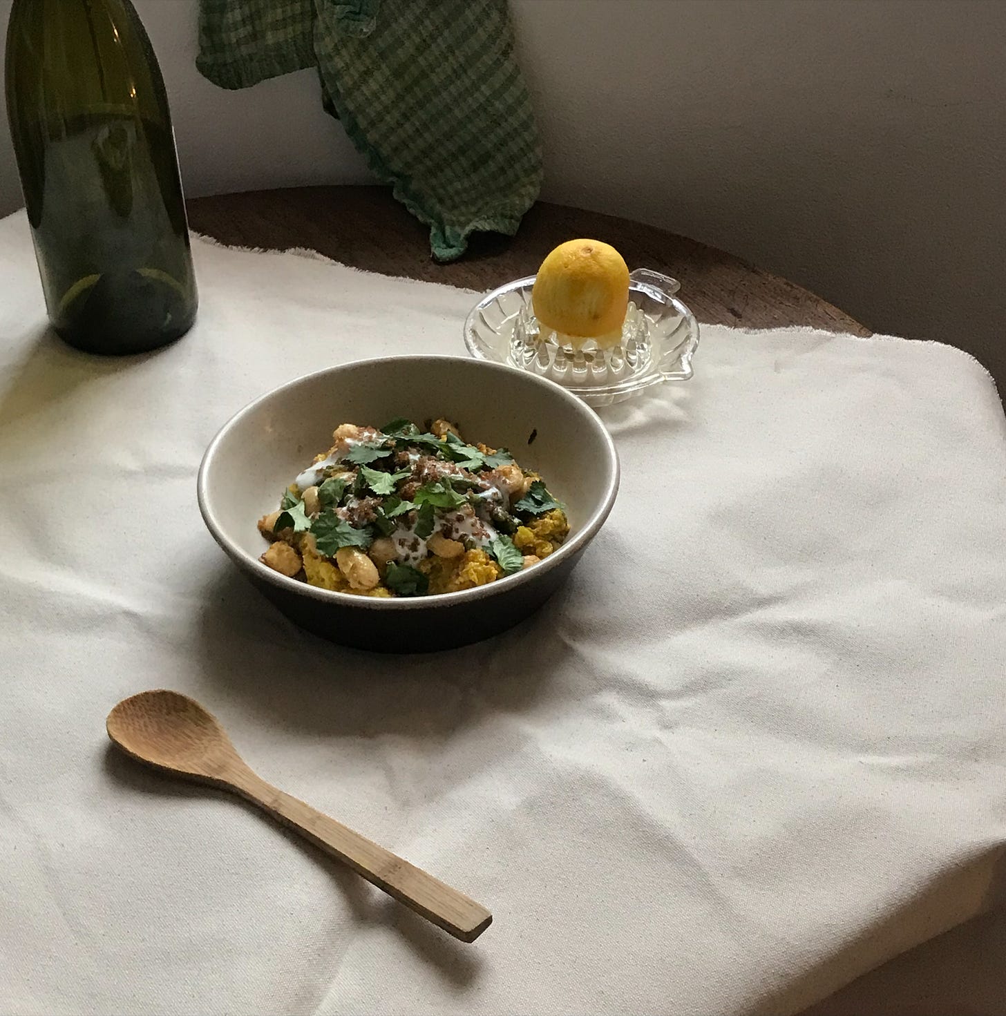 A piece of cream canvas is spread at an angle across a table, with another green fabric hung to the back above it. To the left, a large empty green glass bottle. In the centre, a wide bowl with a mound of yellow butterbeans and green herbs on top, behind which is a glass lemon squeezer with half a lemon atop it. In the foreground, a wooden spoon is placed. 