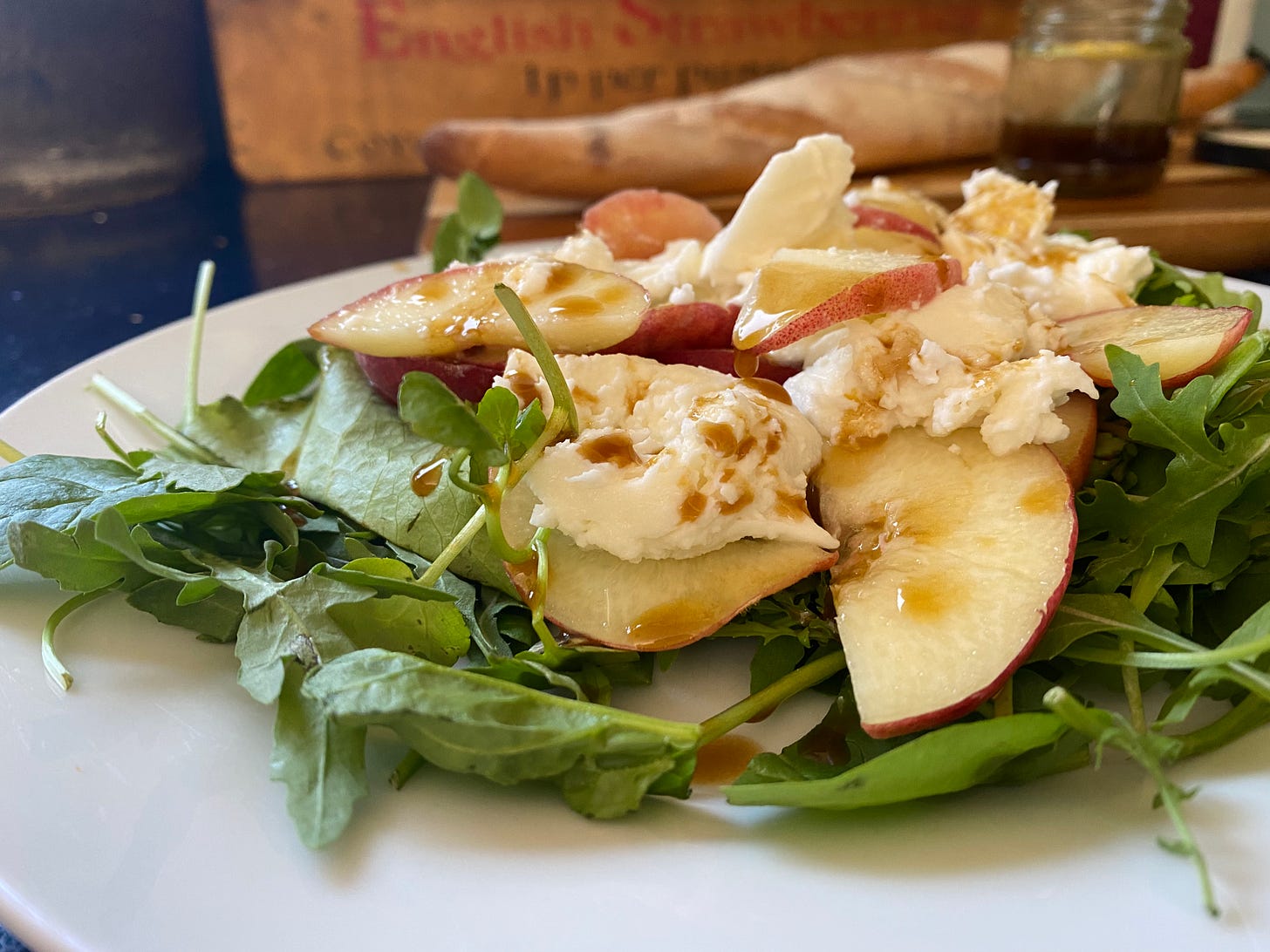 Plate with green salad leaves, topped with peach slices and mozzarella, with a dark brown dressing. In the background a baguette is on a chopping board. 