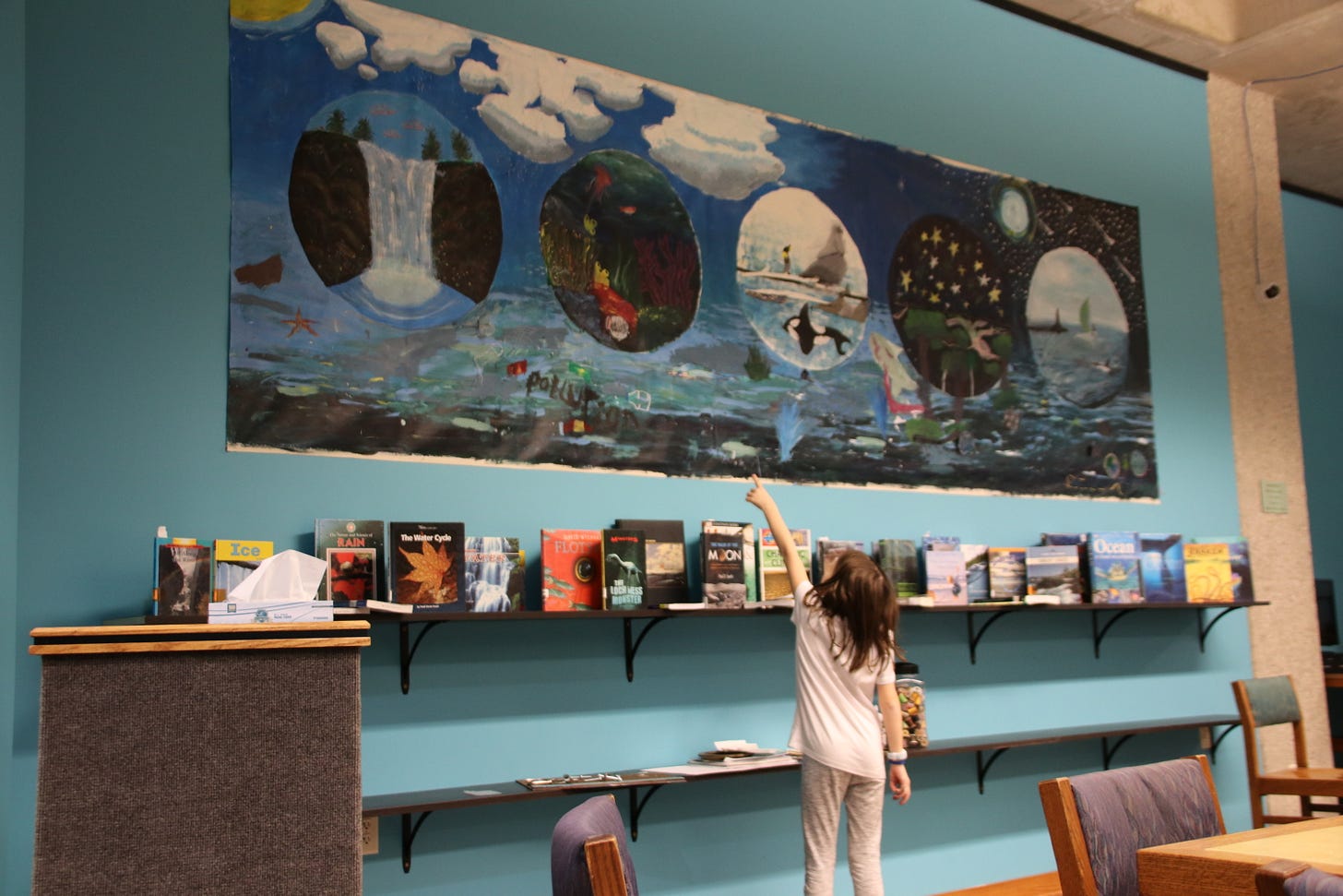 A young child points to a large colorful painting on the wall of a library makerspace.