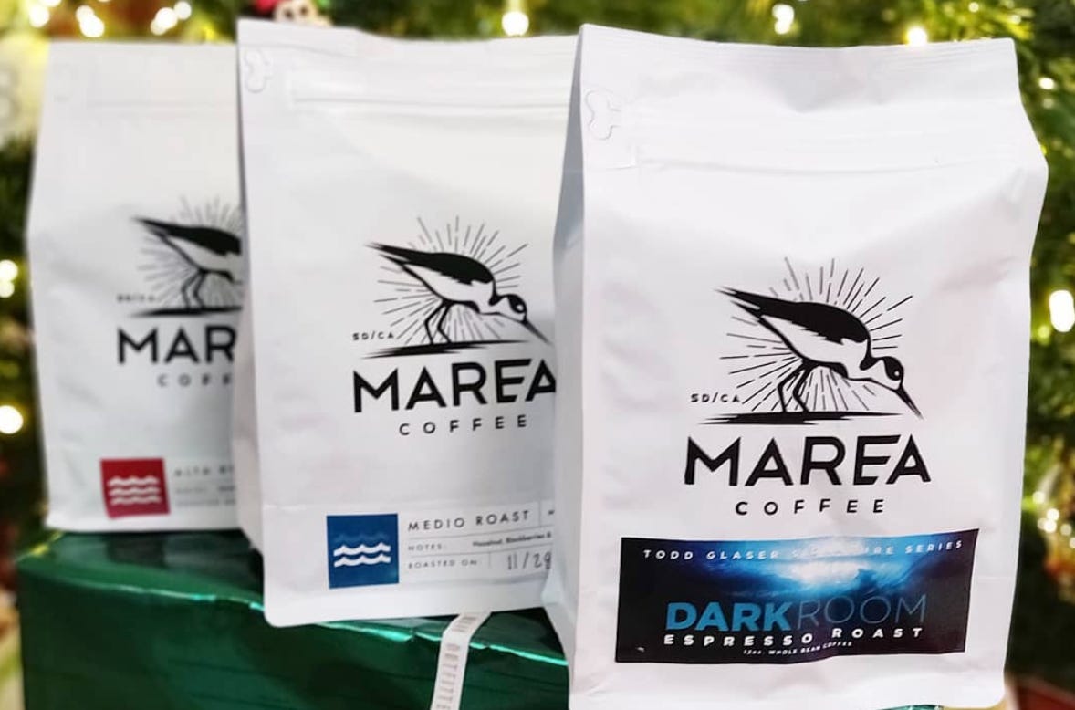 Three white bags of coffee with the Marea Coffee logo of a bird on the beach lined up in front of a Christmas Tree.