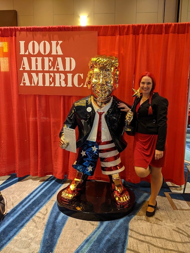 A girl with red hair in a red dress standing next to that gold Trump statue from CPAC dressed in a suit jacket, a tie, and American Flag boxers. A sign in the corner says Look Ahead America. The woman looks super stoked. She is me
