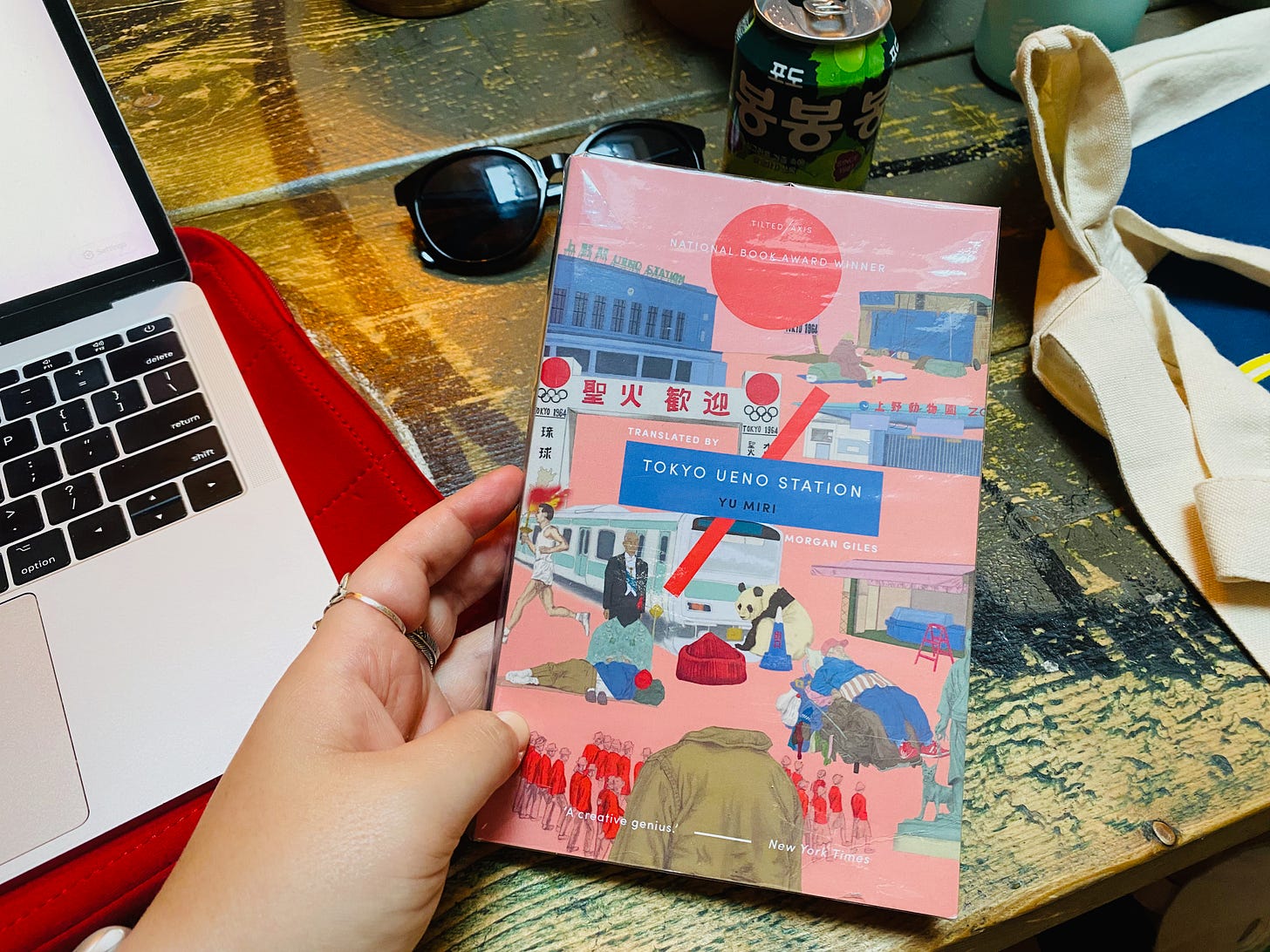 The paperback of 'Tokyo Ueno Station' held up on a wooden table, a laptop to its left and a pair of sunglasses and a Korean drink just above it. Part of a tote bag is seen on the right.