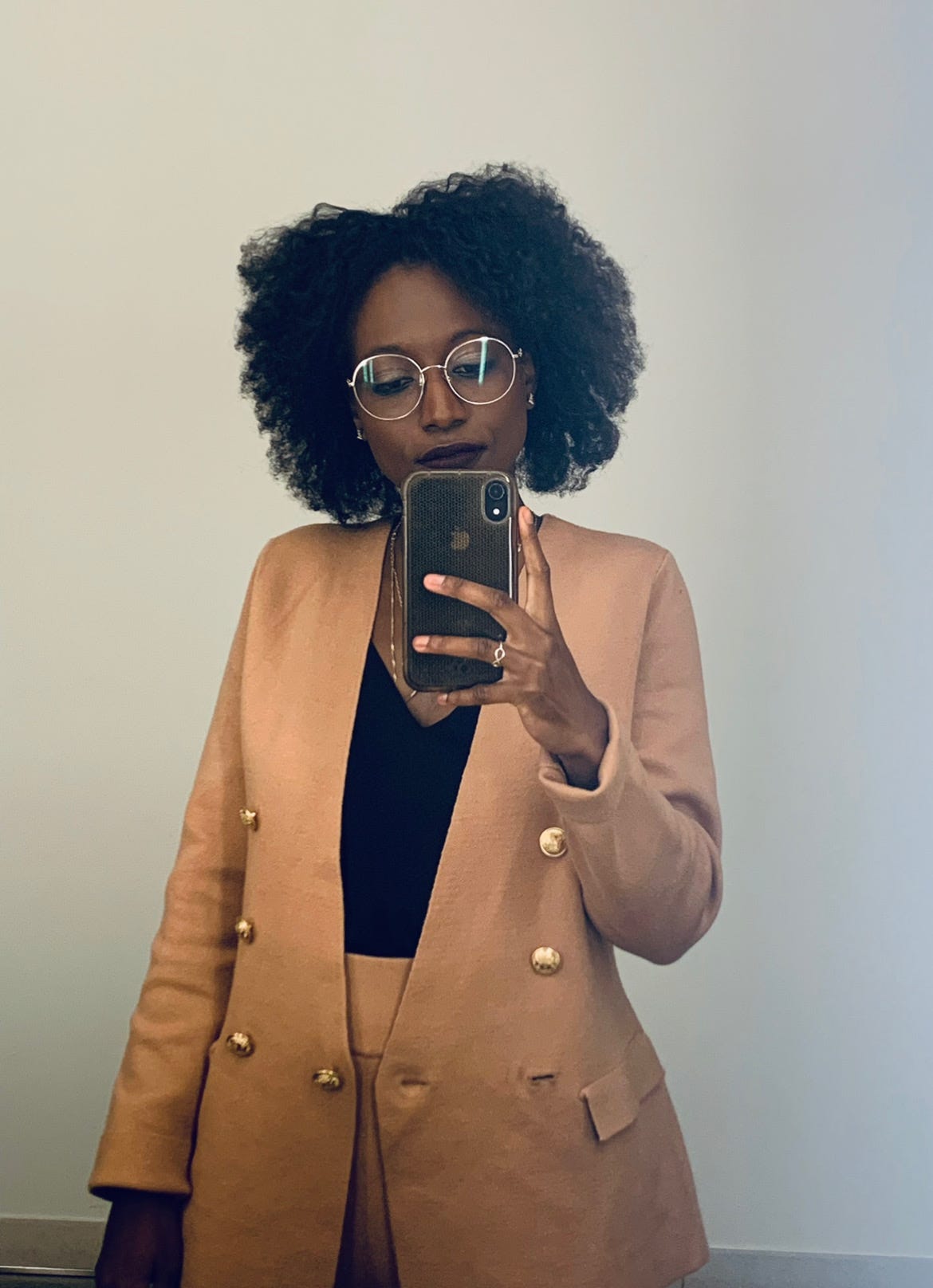 A Black woman taking a picture of herself with an Apple iPhone. She has curly hair and she wears round gold glasses frames, a gold chain necklace, a tan jacket with gold buttons, a black shirt, and tan pants.