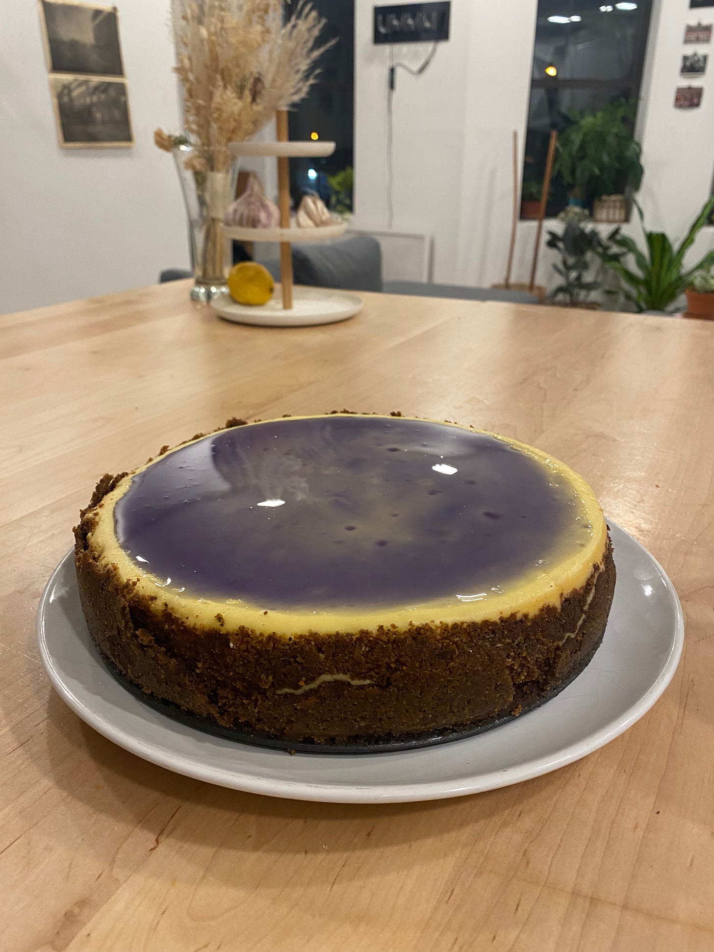 A cream cheesecake with a blue glossy syrup and brown crust sits on a white plate and wood kitchen island.