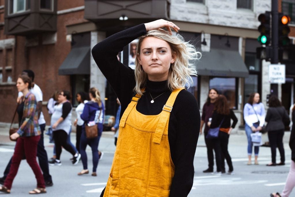 person standing in a city street wearing yellow overalls, with hand on head and a confused expression on their face. people walk past in the background