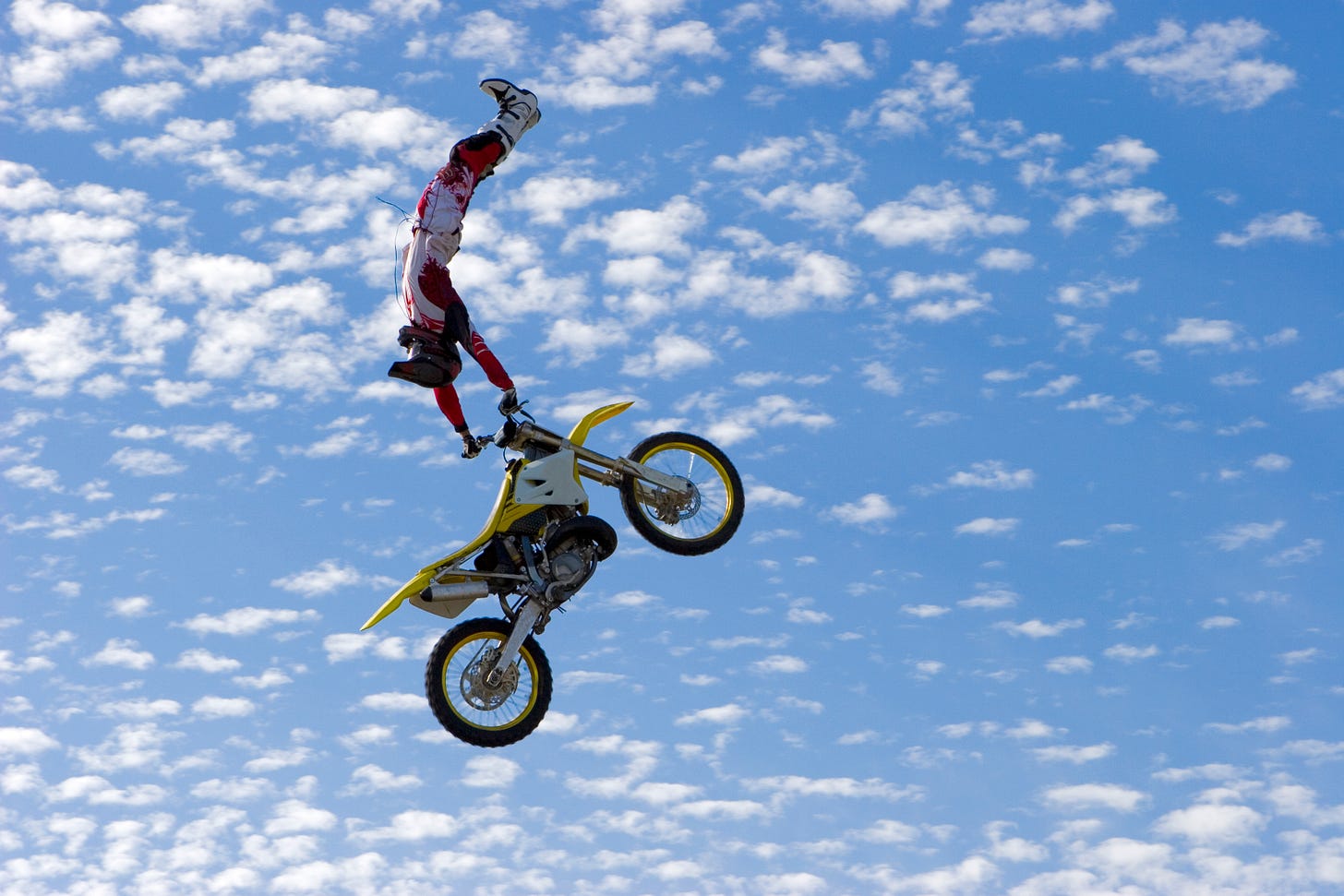 Motocross aerialist completely upside down holding the handlebars of a bike in a blue but cloudy filled sky, the bike wheels down