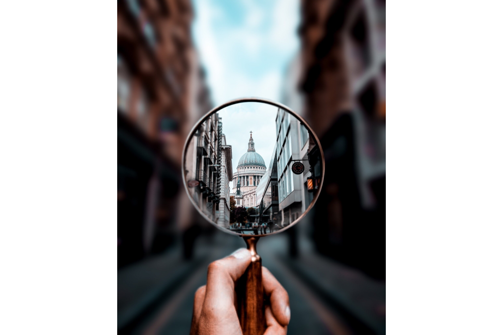 Hand holding up a magnifying glass showing a building and blurry background.