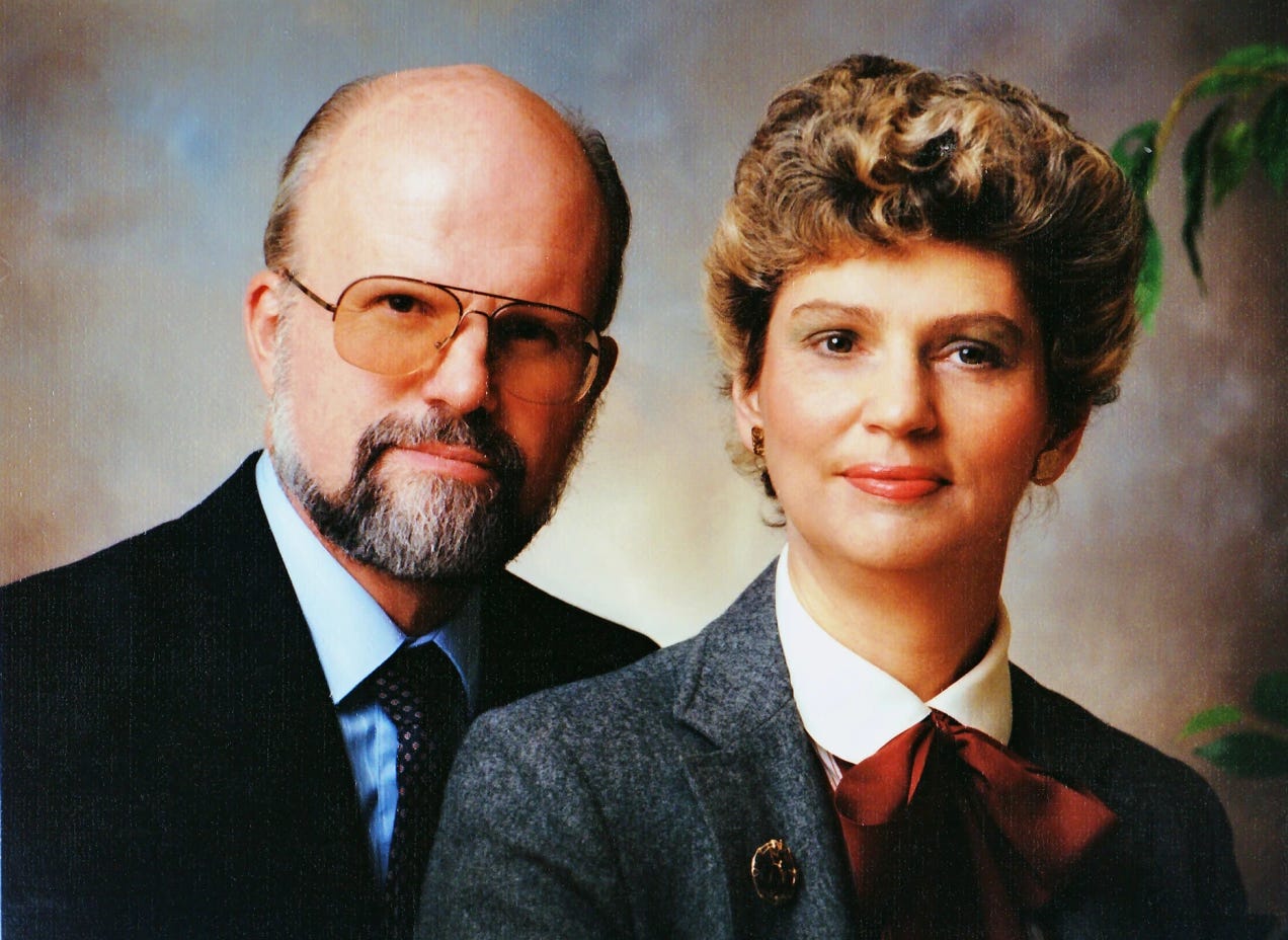 Formal portrait of a couple in dress suits looking at the camera, both smiling slightly. The man has glasses, a graying beard, and a mostly bald head. the woman is wearing earrings, light make-up, and has short, curly, frosted hair