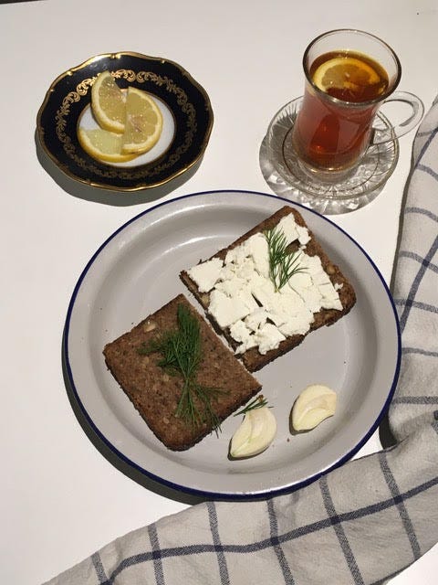 A photo from above of a plate with two slices of rye bread, one covered with crumbly white cheese, some dill. A split garlic clove beside. Around, a tea glass and saucer with an amber liquid in, and another saucer with lemon slices. 