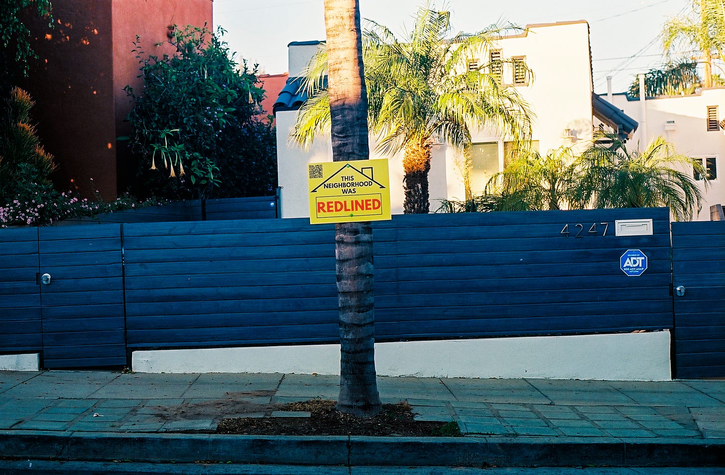 Photograph of a black wooden gentrifence in front of a renovated apartment complex with one of my signs nailed to a palm tree in the center of the frame.