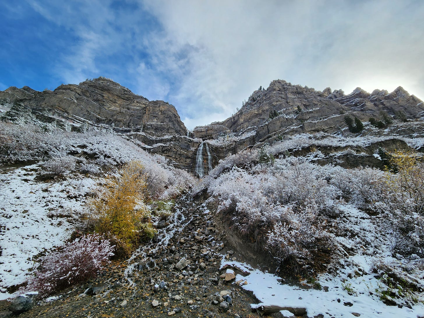 thin waterfall amid mountains with light snow on rocks, ground, and brush