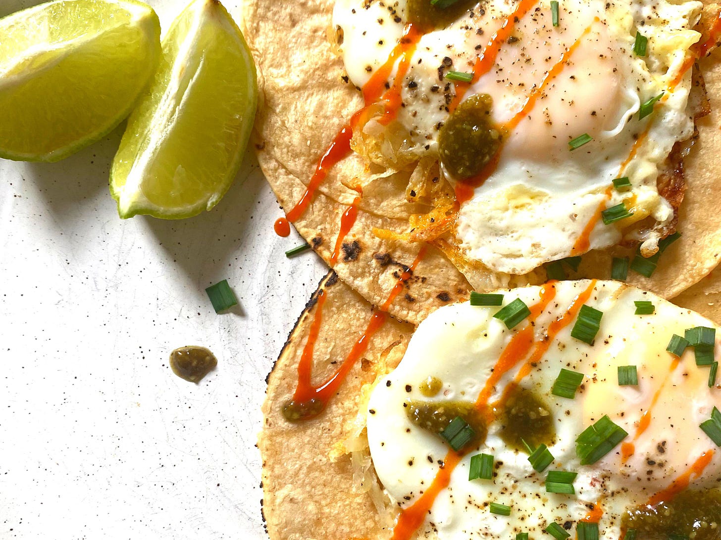 Two tortillas, topped with potato, eggs, hot sauce and green vegetable. Two lime wedges are placed next to it.