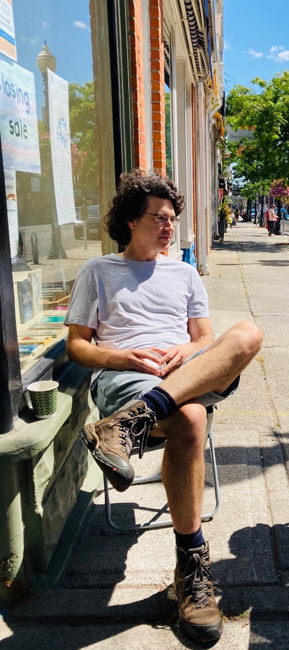 Bookseller James McDonald sitting outside The Printed Word bookshop. Closing sale sign in window behind him.