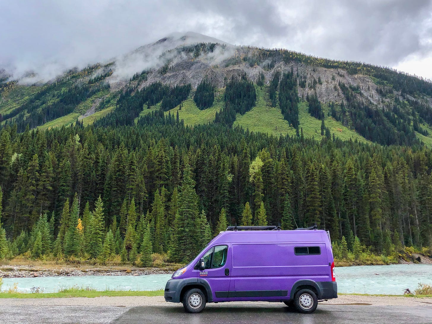 Purple van parked by a mountain and river.