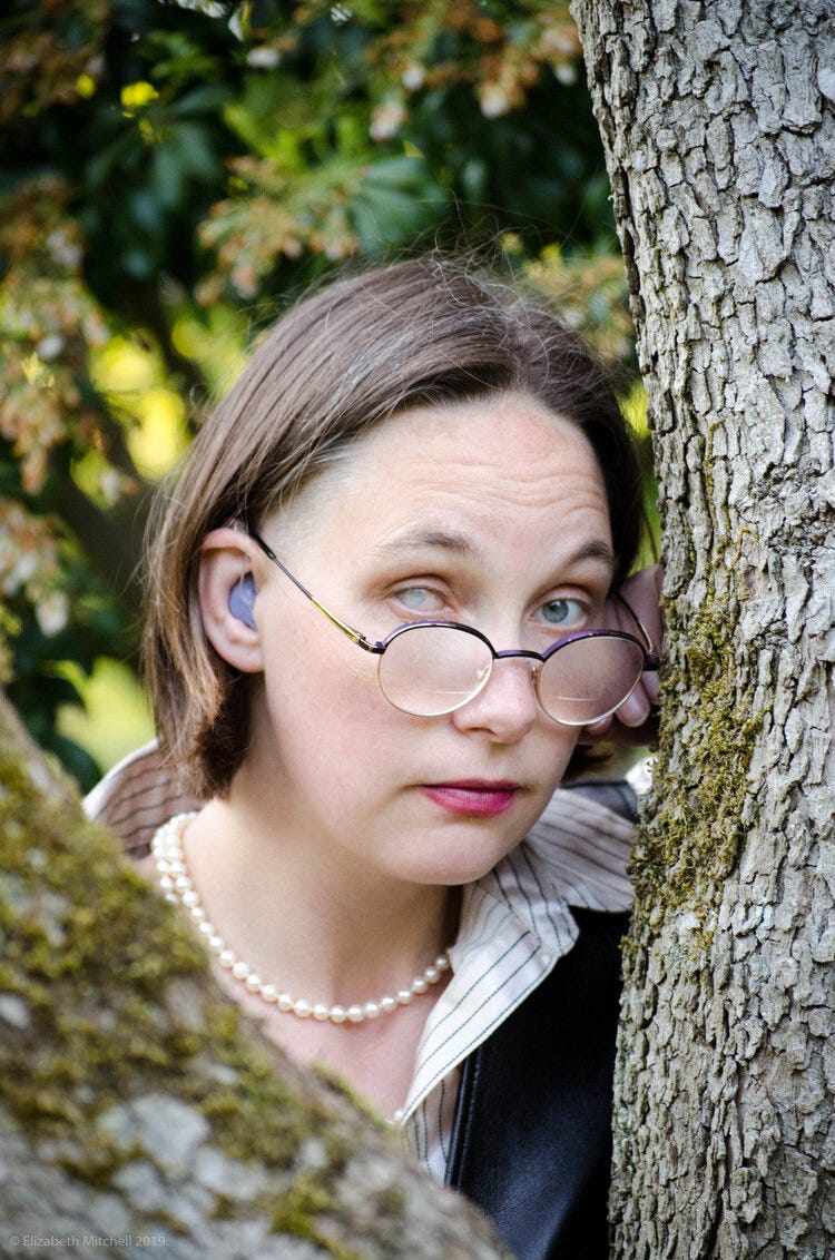 A white woman with short hair and an occluded cataract on her right eye wearing purple hearing aids and a pearl necklace, looking with raised eyebrows at camera between trees. Photo credit: Lis Mitchell, 2021