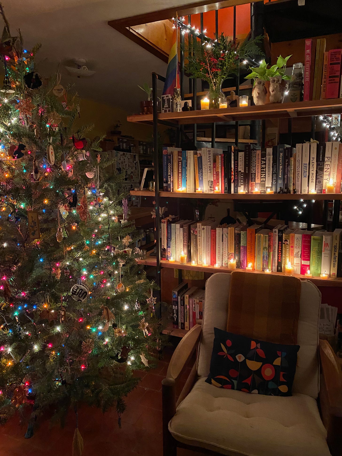 A large open bookcase full of cookbooks, with rows of lit candles in front of the books. There are lights on the spiral staircase behind the bookcase, and brightly lit and decorated Christmas tree in front. A armchair with a small throw pillow sits next to the Christmas tree.