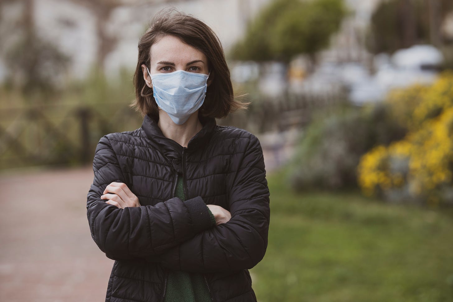 woman in black puffy jacket standing outdoors with arms crossed wearing a medical mask staring straight at the camera
