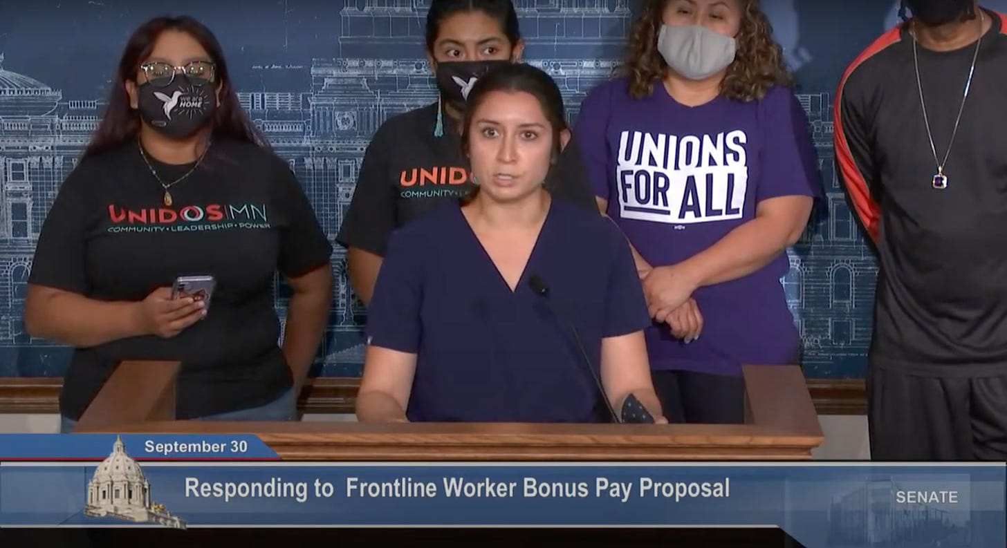 A white woman with brown hair in navy scrubs stands behind a podium, behind her people in graphic tees reading Unidos MN and Union for All