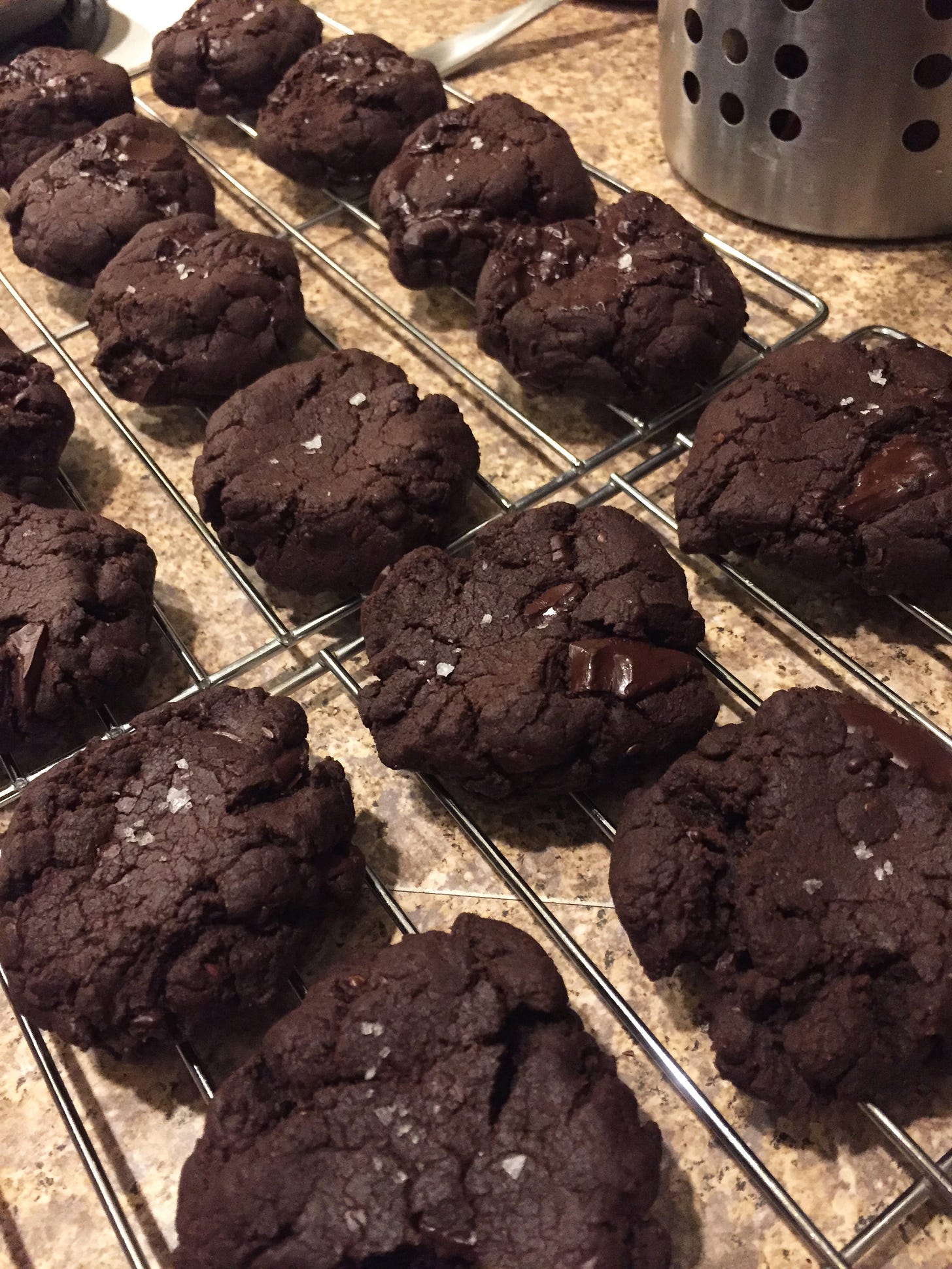On a cooling rack, lines of thick, dark brown double chocolate cookies with salt on top.