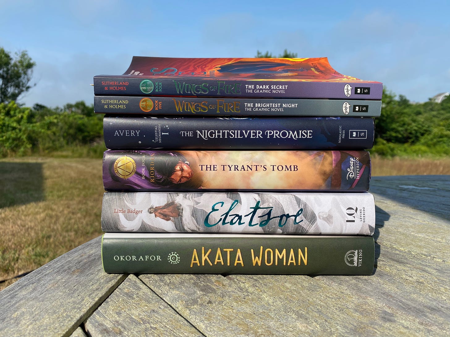 A stack of books on a picnic table in a sunny yard. The books arel Akata Woman, Elatsoe, The Tyrant’s Tomb, The Nightsilver Promise, and two volumes of The Wings of Fire graphic novels.