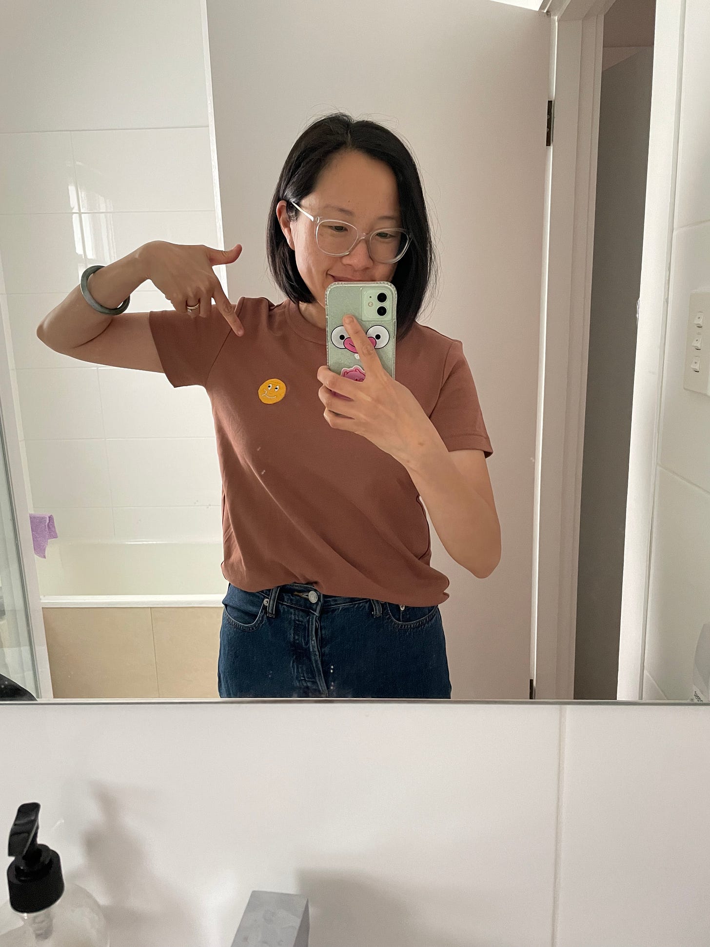 Selfie in bathroom mirror wearing a brown shirt with a hand embroidered chewing smiley face on it.