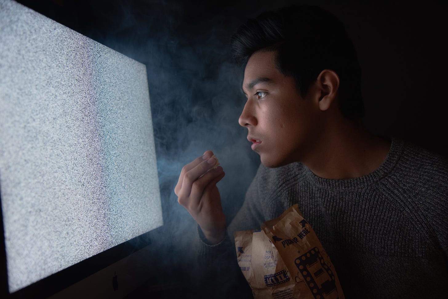 A man stares at a TV screen in a dark room while eating popcorn.
