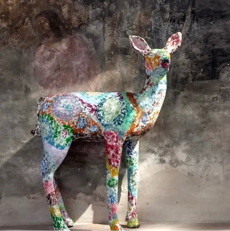 A sculpture of a doe, she is multicolored and appears to be made out of paper maché. Light is streaming onto her and she looks really cute.