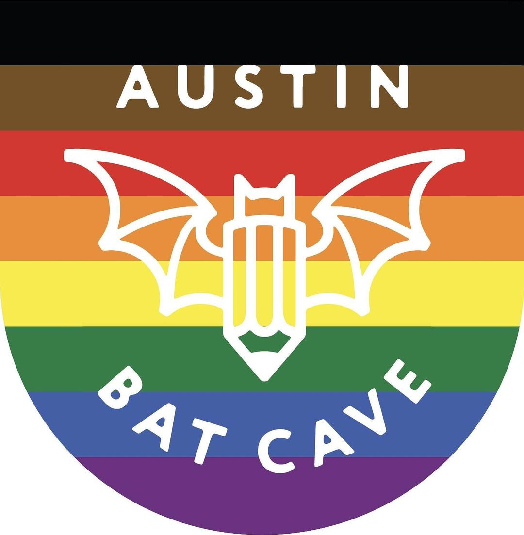 The Austin Bat Cave logo, a bat with the body of a pencil, lays atop black, brown, red, orange, yellow, green, blue, and purple stripes to celebrate Pride.  