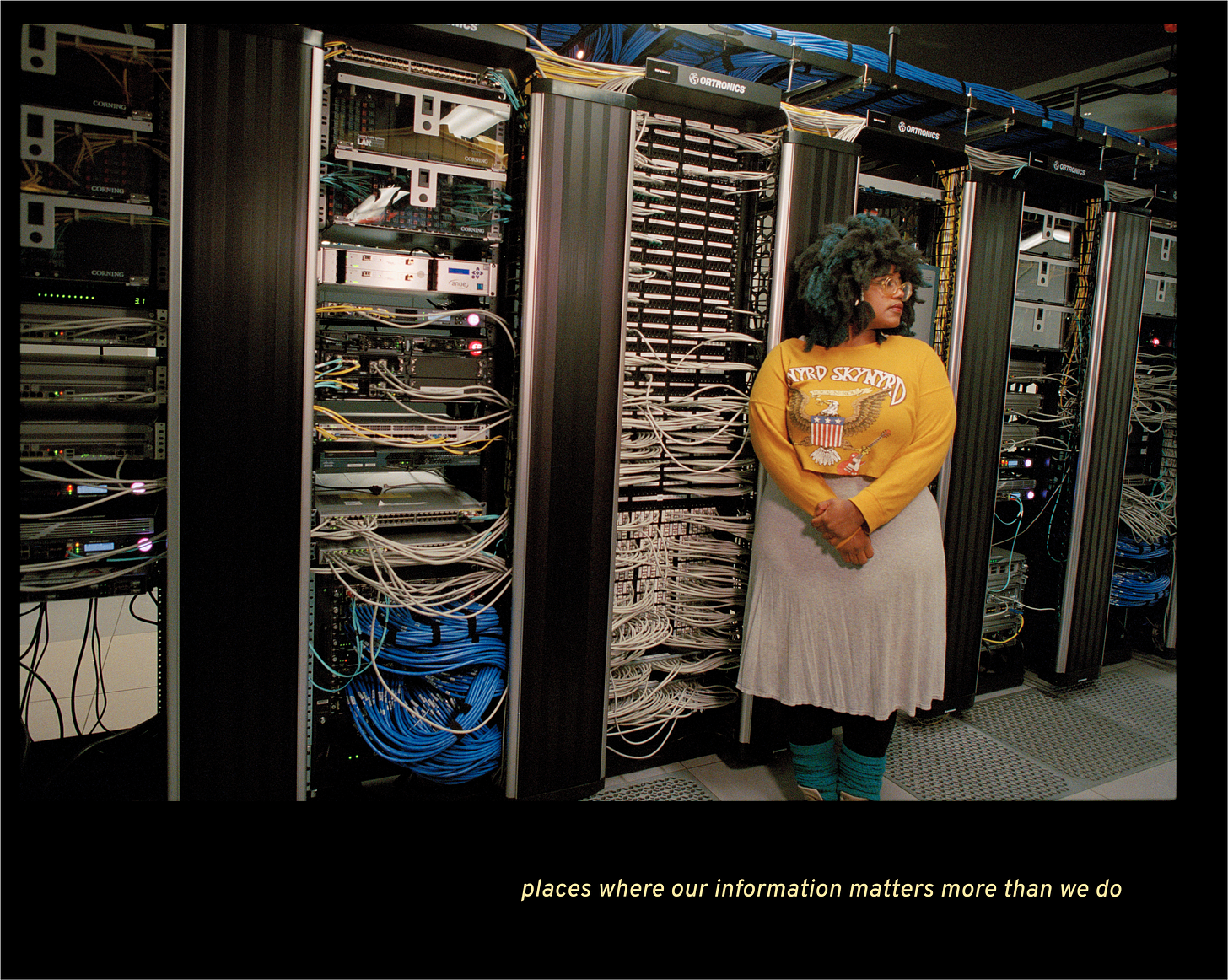 Image of a Black woman standing in front of a wall of computers, cables, and devices. She looks away from the camera.