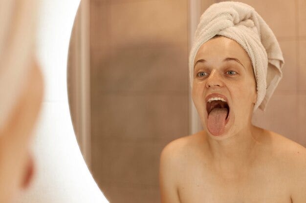 Foto interna de feamle ensolarado posando no banheiro depois de tomar banho, olhando para seu reflexo no espelho com a língua de fora, olha para a língua dela, procedimentos higiênicos. Foto gratuita