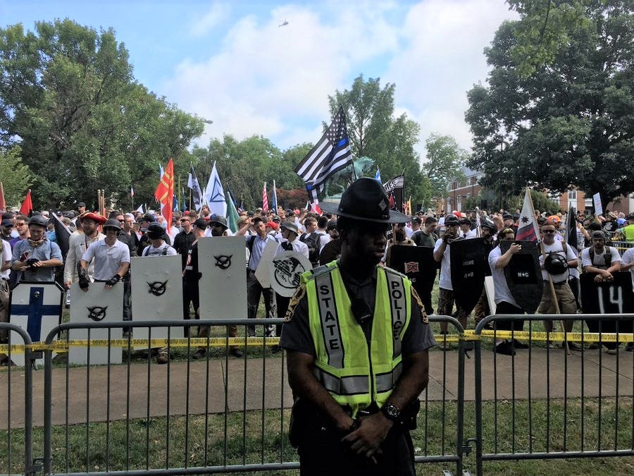 A uniformed State Police officer in a reflective vest stands with his back to a crowd displaying a Thin Blue Line flag and various white supremacist imagery and slogans at the 2017 Unite the Right rally in Charlottesville, VA. The officer is a Black man.