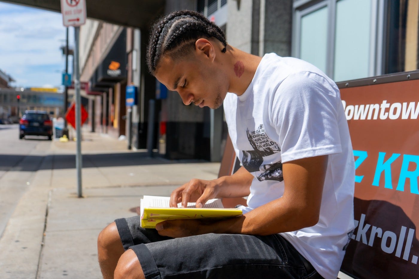 A casually dressed young man reads a book on a city bench. He is engrossed in the reading. Urban landscape of signs, buildings, and cars are in the background.
