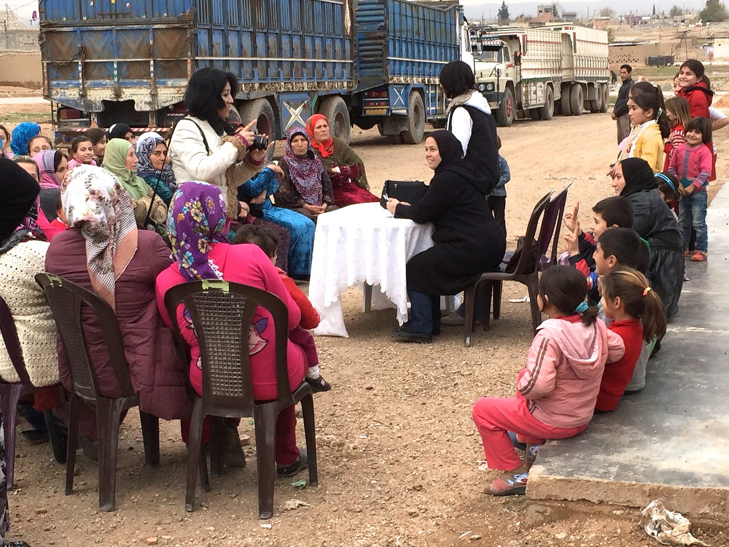Kurdish women and children sit in outdoor chairs,, gathered around a local representative. 