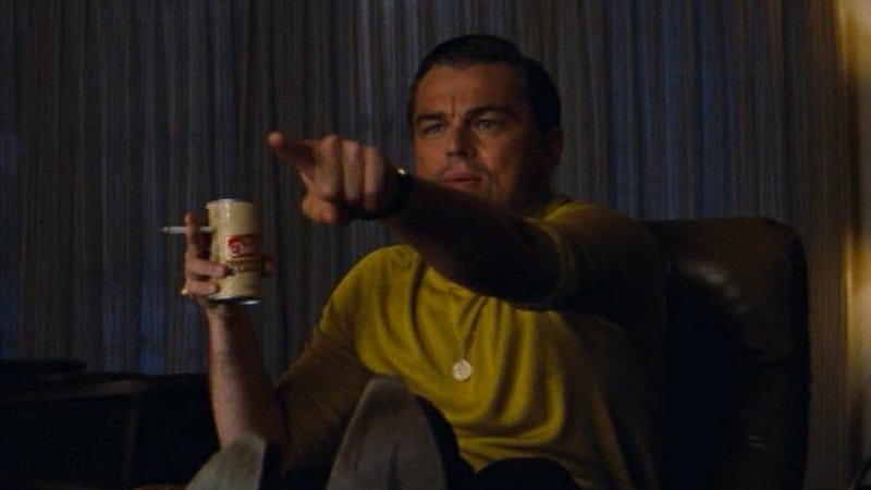leonardo dicaprio as rick dalton in the film once upon a time in hollywood sitting on a couch in a yellow sweater and holding a beer and pointing his finger at the television screen.LoadOfBarnacles07
