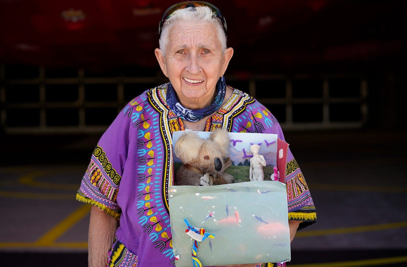 An older, queer person wearing a purple Dashiki holds their diorama-like artwork.