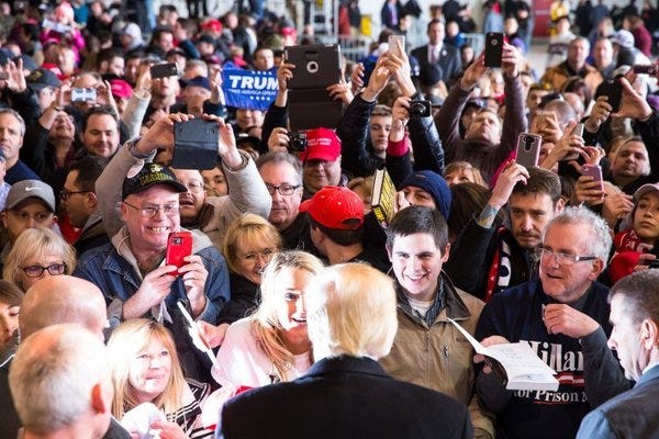 A mass of delighted white people crowd around Donald Trump, seen from behind.