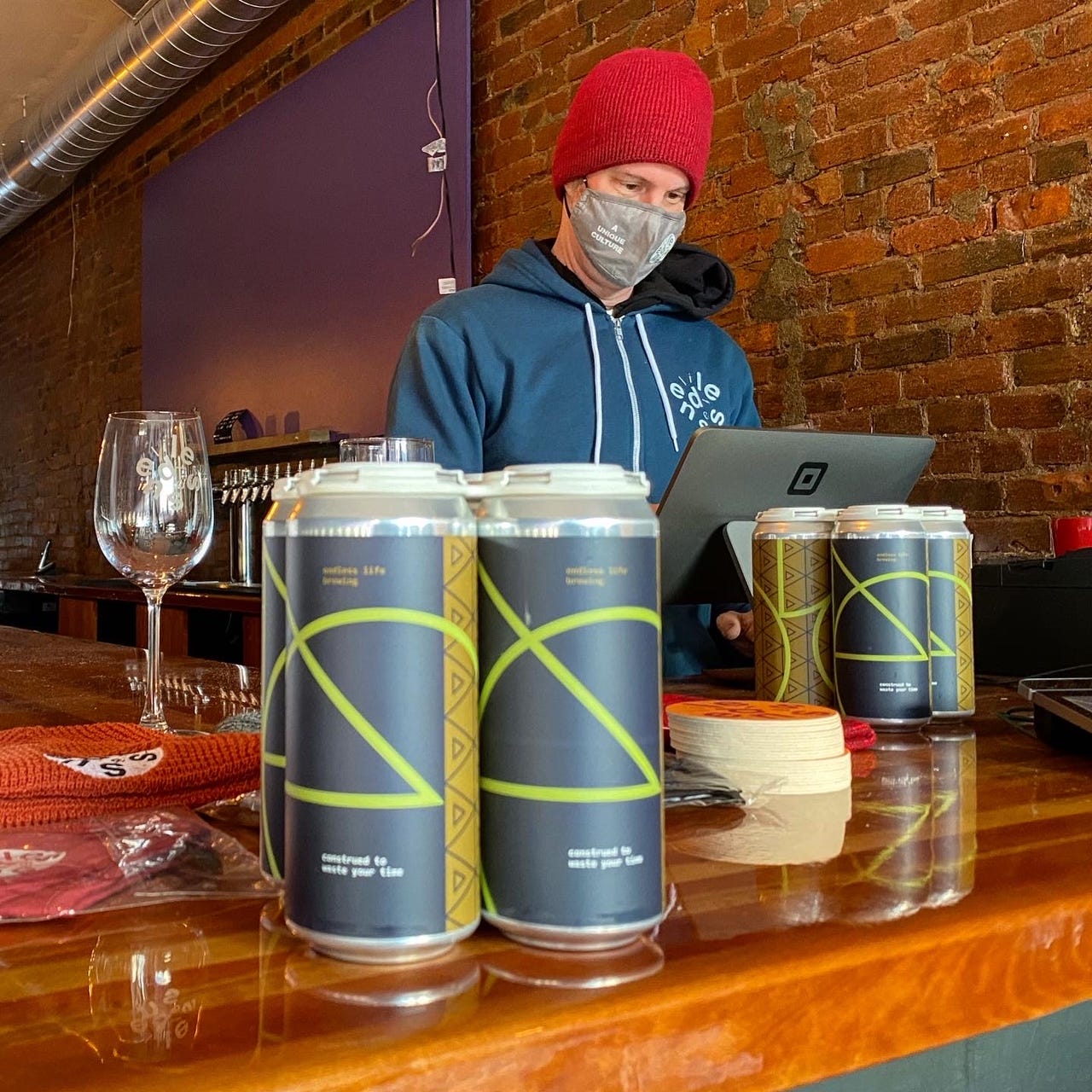 A four-pack of cans sits on counter while a masked brewer uses cash register