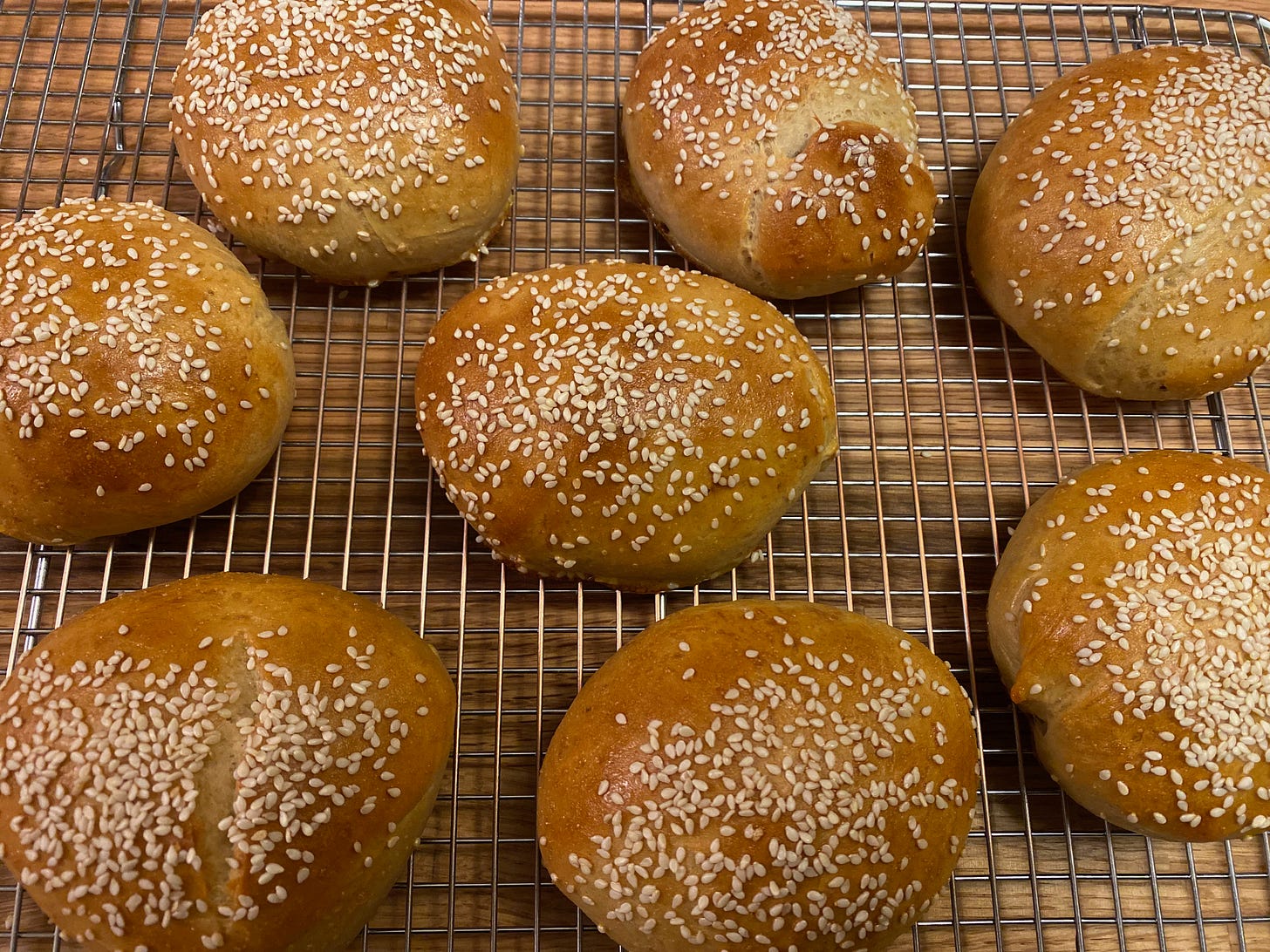 Eight round, golden-brown buns covered in sesame seeds sit on a metal cooling rack.