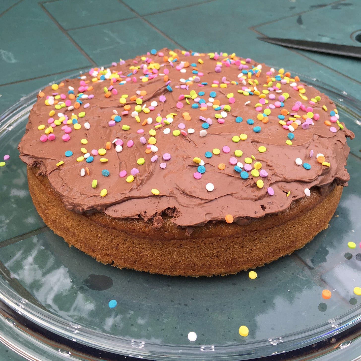 On a green patio table, golden yellow cake with a thick layer of chocolate mousse spread over the top, and large round sprinkles dusted over it. 
