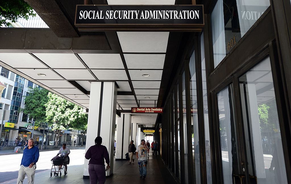 People walk on the street outside a Social Security Administration office, indicated by a large sign above the street, on a sunny day.