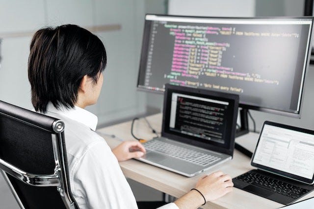shot of person from behind. they are wearing white, seated at a desk, coding, with three screens in front of them