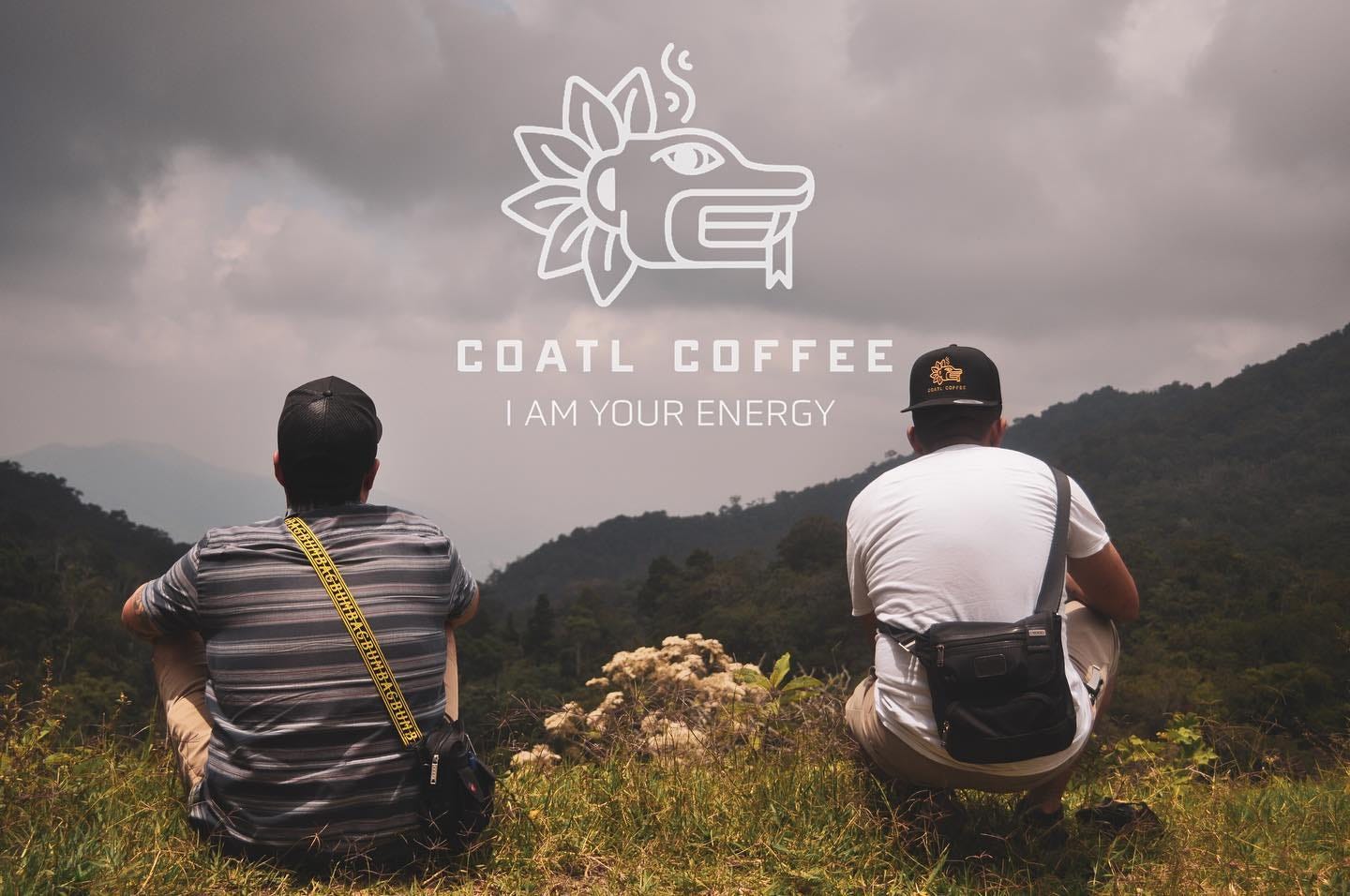 A photo of two men crouching down on a wild, grassy patch looking out over mountain forests under a cloudy sky. The Coatl Coffee logo, a line drawing of a feathered serpent head based on Aztec myth along with the phrase "I am your energy" is imposed over the clouds.