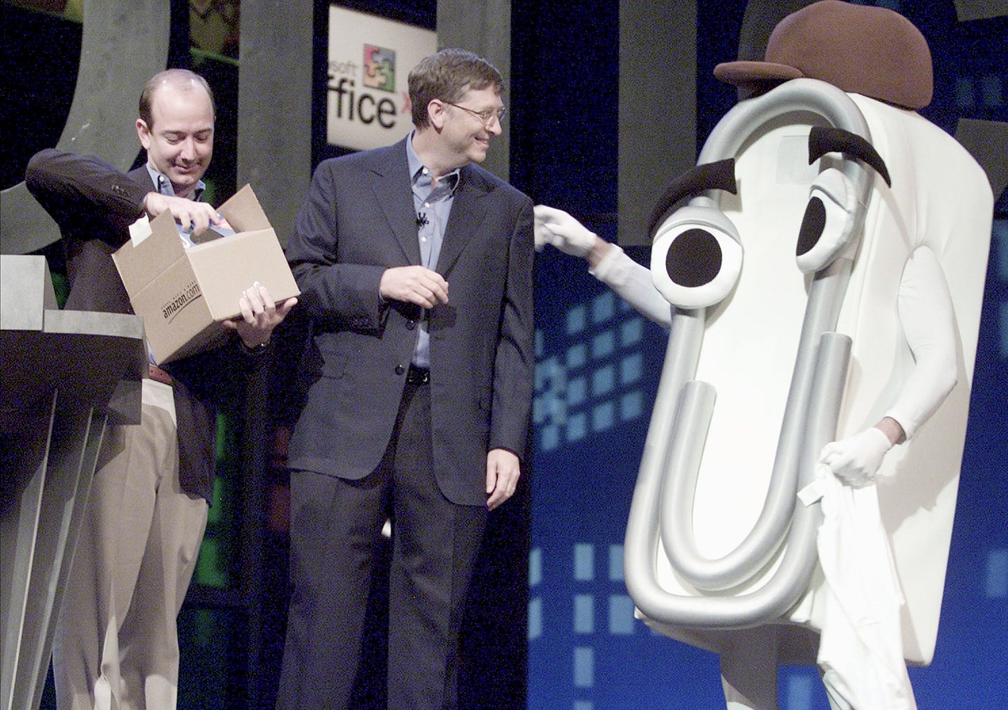 Photo on stage of Jeff Bezos opening an AMazon box with Bill Gates interacting with a person dressed as Clippy.