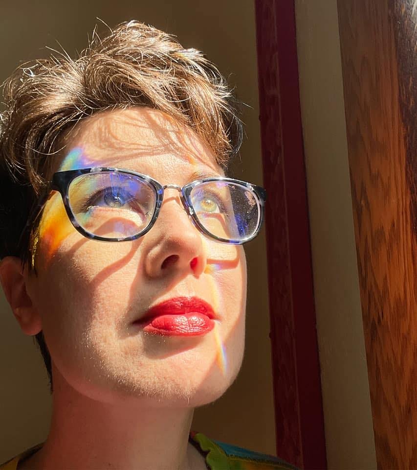Misty Gedlinske is a white woman with short, wavy brown hair and glasses. She is pictured here from the neck up, looking upward into the sun through a window with rainbow prisms on the left side of her face.