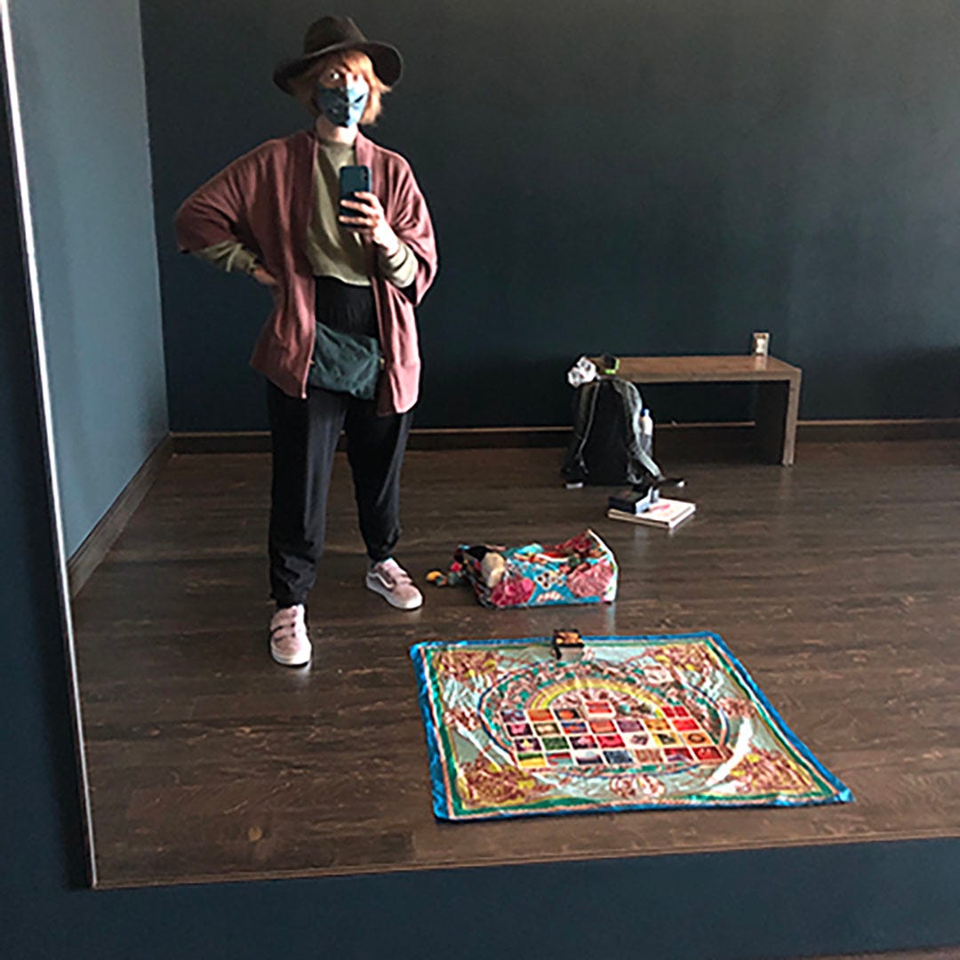 A photo of ilyse taking a picture of herself in the mirror including a full spread of cards on a scarf on a wooden floor in the SFO Yoga Room.