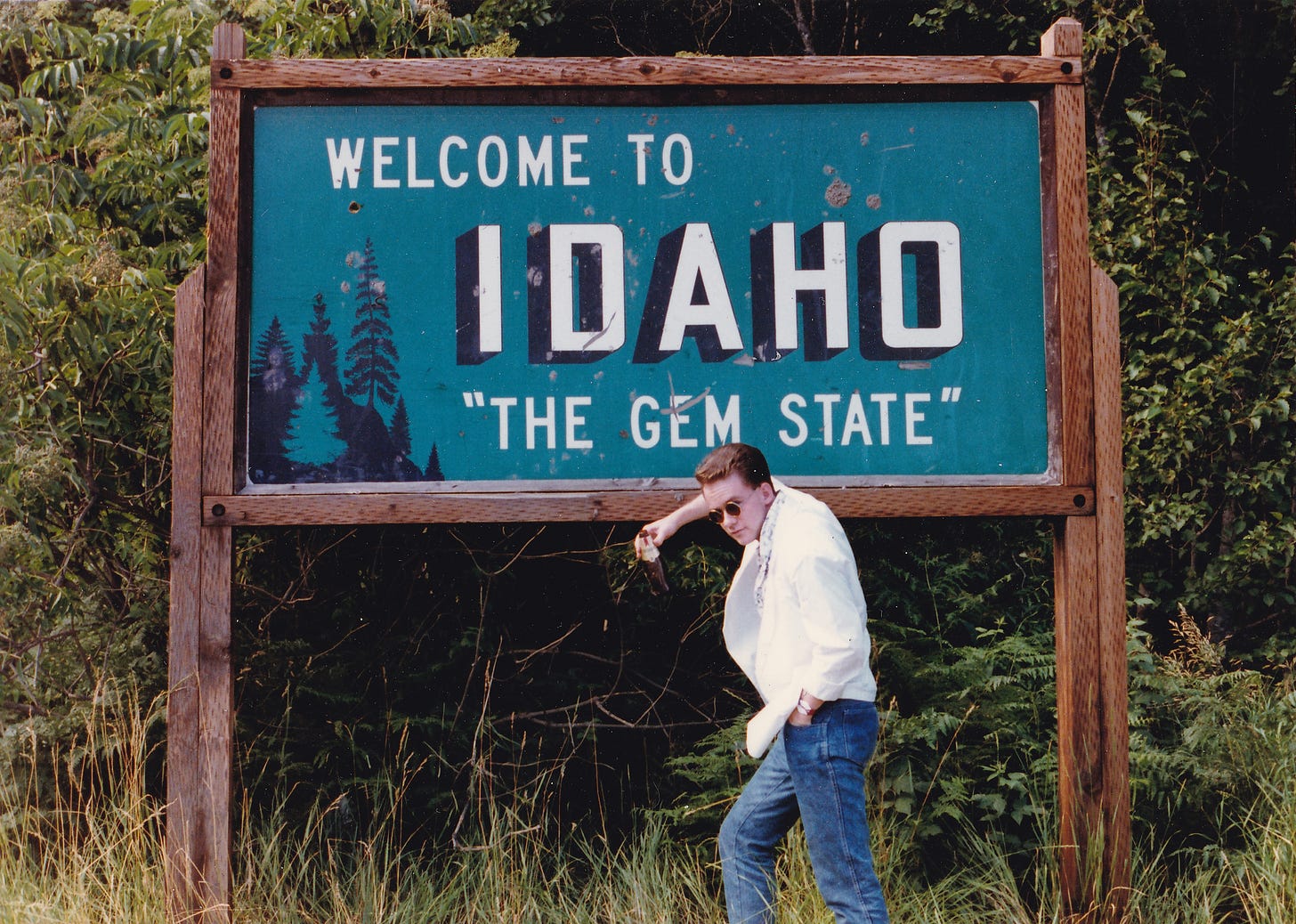A young white man wearing sunglasses, a white blazer, and jeans, poses insolently leaning against a huge "Welcome to Idaho, The Gem State" sign, against a background of trees and undergrowth. He holds a beer bottle.