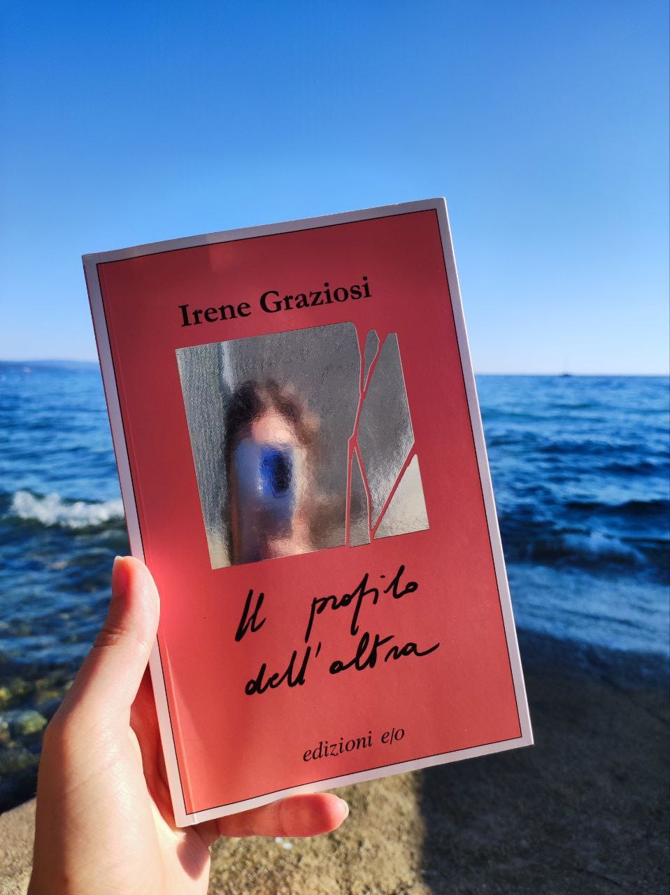 Sullo sfondo il mare, al centro una mano tiene un libro di colore rosa. Il titolo è "Il profilo dell'altra" e l'autrice è Irene Graziosi. Sulla copertina c'è un piccolo specchio.