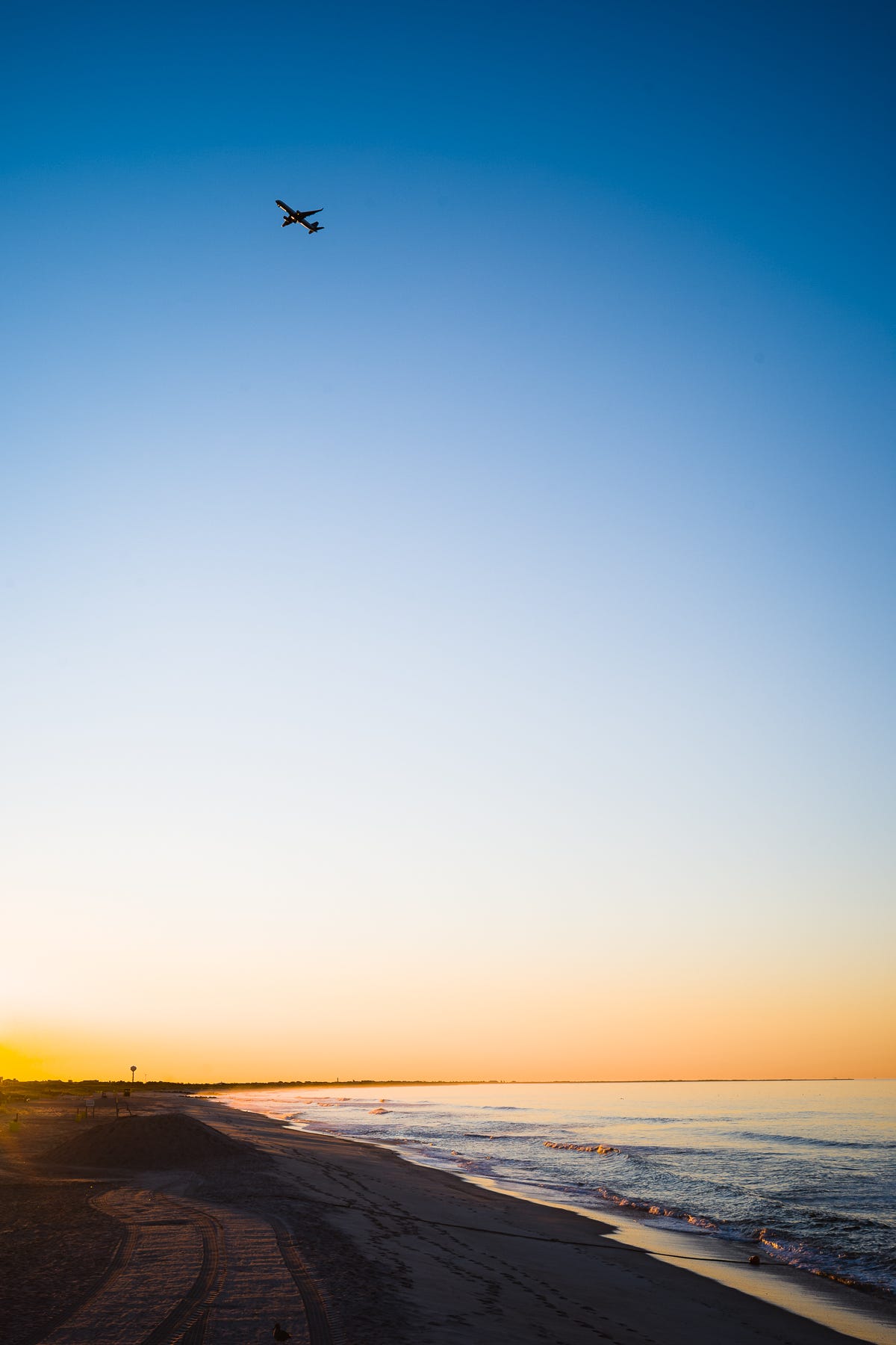 A plane flies over a beach at sunrise in a bright blue and orange sky as waves break against the sand